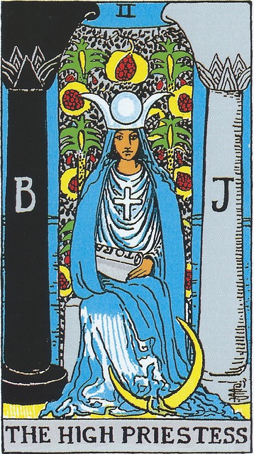 Tarot Card - The High Priestess 
A female figure is seated on a throne, on her head a horned diadem representing the phases of the moon.  She wears white robes with a cloak of blue, and a cross on her chest.   In her lap, and partially concealed by her cloak, lies a partially unrolled scroll with the letters T O R A visible.  Is this the Torah as many believe?  On either side of her are two pillars, one black, one white.  Behind her is a curtain, or veil, with pomegranates.  At her feet is a crescent moon. Glimpses beyond the veil reveal the sea. 