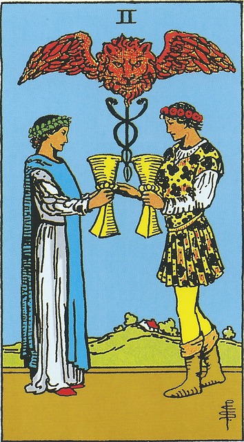 The Two of Cups:  Two figures, a male and a female.   The female wears long white robes with a blue mantle, reminiscent of the High Priestess, with a laurel wreath on her head. The male wears a tunic, leggings, and boots, colored and reminiscent of the fool, and is crowned with a floral wreath.  They each hold out a golden vessel to the other, from which a caduceus – a symbol of healing – rises.  The male is also extending a hand to the female.  Overlooking all is a red lion’s head sprouting wings.  In the distant background we see the red roof of a house that is surrounded by trees and perched on a gentle slope.  